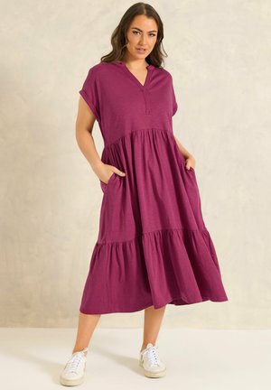 Femme portant une robe midi violette ample à volants et manches courtes avec poches, associée à des baskets blanches, debout devant un fond uni.