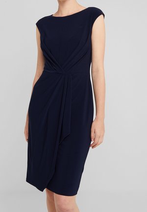 Frau trägt ein ärmelloses knielanges marineblaues Kleid mit gerafften Stoffdetails an der Taille und einem asymmetrischen Frontdrapé.