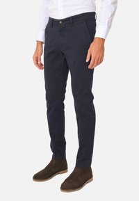 Chino slim-fit blu navy in cotone con frontale piatto, tasche laterali e chiusura a bottone, indossati con stivali alla caviglia in suede marrone.
