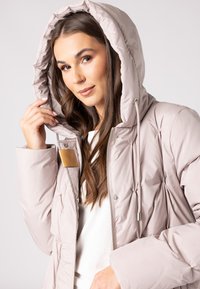 Doudoune rose clair avec capuche, dotée d'une fermeture éclair, d'une texture matelassée et d'un accent métallique près de la fermeture.