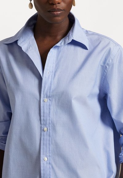 Femme portant une chemise bleu clair à boutons avec manches retroussées et grandes boucles d’oreilles ovales, montrée des épaules jusqu’au milieu de la poitrine.