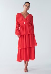 Abito maxi rosso a strati realizzato in tessuto leggero. Caratterizzato da maniche lunghe, scollo a V e vita elastica con un accento metallico.