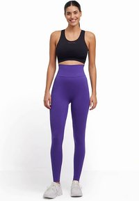 Zwart crop top met een klein logo, hoge paarse leggings met een getextureerd patroon, gecombineerd met witte sportschoenen.