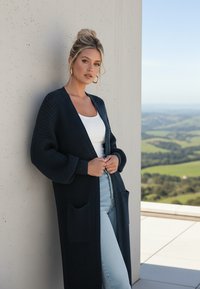 Długi, granatowy kardigan dzianinowy o teksturowanym wykończeniu, z otwartym przodem, dwiema przednimi kieszeniami i ściągaczami na rękawach, noszony na białą bluzkę i dżinsy.