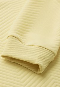 Pullover giallo con polsini a costine e motivo zigzag testurizzato sul tessuto. Materiale morbido, primo piano del dettaglio della manica.