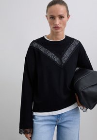 Femme aux cheveux tirés en arrière portant un pull noir avec un détail en dentelle en V, un jean bleu clair, et portant un sac en cuir noir sous le bras.