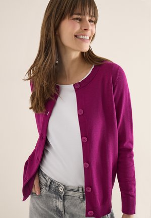 Donna sorridente che indossa un cardigan viola acceso con bottoni sopra una camicia bianca e jeans grigi, mani nelle tasche, davanti a uno sfondo neutro.