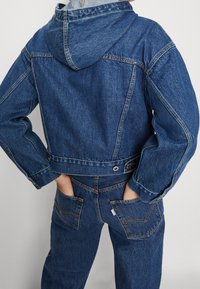 Džínová bunda s kapuc í v tmavě modré barvě. Má prošité švy, přední kapsy a zkrácený střih s knoflíkovým zapínáním.