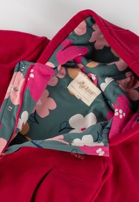 Giacca in pile rossa con fodera stampata floreale in verde, caratterizzata da fiori rosa e bianchi, chiusura a scatto e etichetta del marchio all'interno.