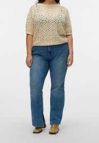 Donna che indossa un top a maniche corte lavorato all'uncinetto beige, jeans dritti blu e scarpe beige con punta quadrata, in piedi davanti a uno sfondo semplice.