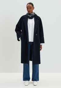 Marineblauer Wollmantel mit weiten Ärmeln und großem Kragen. Kombiniert mit einem weißen Hemd, dunkelblauen Jeans und weißen Sneakers.