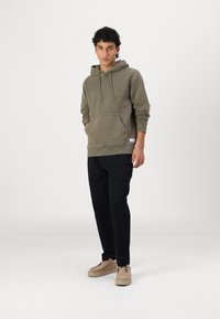 Olivgrüner Hoodie mit einer Vordertasche und verstellbarem Kordelzug, kombiniert mit schwarzen Hosen und hellbeigen Schuhen, vor einem weißen Hintergrund stehend.