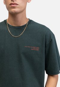 T-shirt en coton vert foncé à manches courtes et col rond. Présente un texte rouge sur le devant : "YEUX GRAND OUVERTS QUI S'EN SOUCIE REDEFINI REBELLE."