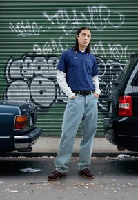 Plava karirana polo majica preko bijele majice s dugim rukavima, izblijedjele plave traperice i smeđe cipele s vezicama, na pozadini s grafitima.