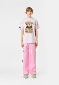 Camiseta oversized de color rosa claro con un gráfico de un oso de peluche, combinada con pantalones jogger rosa a juego. Zapatillas grises texturizadas sin cordones.