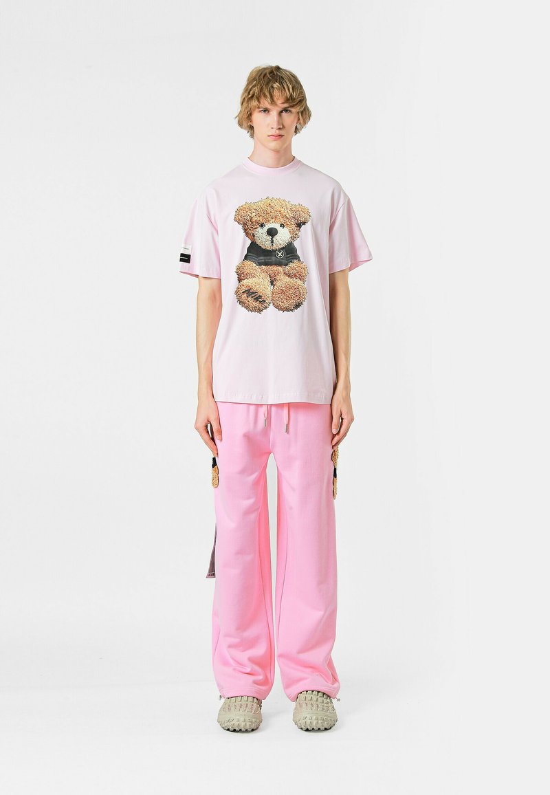 Camiseta oversized de color rosa claro con un gráfico de un oso de peluche, combinada con pantalones jogger rosa a juego. Zapatillas grises texturizadas sin cordones.