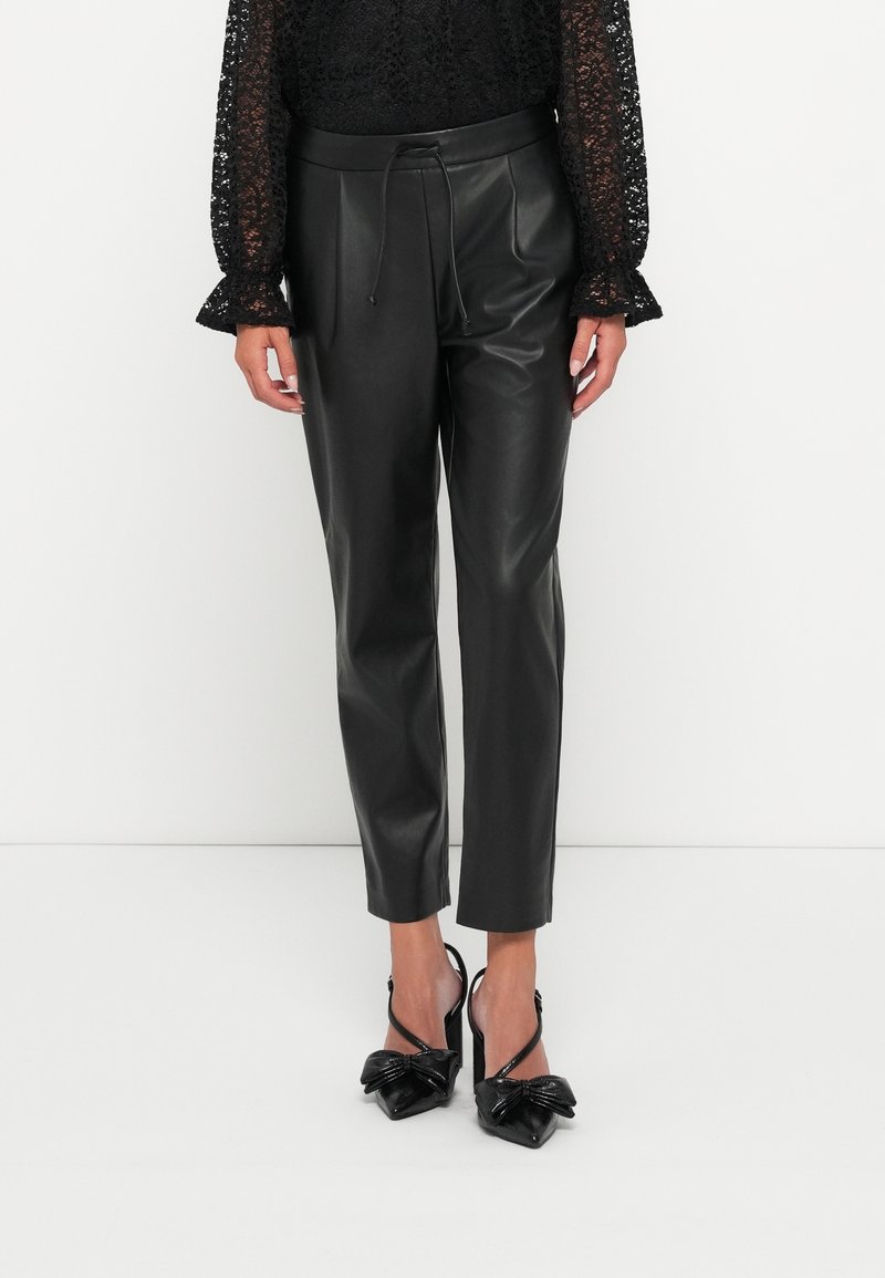Pantalon noir en simili cuir taille haute avec cordon, associé à un chemisier en dentelle noire et des escarpins noirs à bout pointu avec détails de nœud.