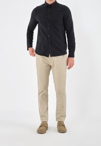 Camisa negra de manga larga con cuello, pantalones beiges y zapatos marrones de slip-on. Tela suave, cierre de botones y diseño ajustado.