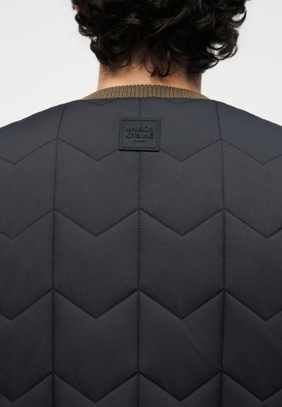 Άτομο που φαίνεται από πίσω φορώντας μαύρο καπιτονέ μπουφάν με ετικέτα Maison Kitsuné Paris κάτω από τον λαιμό και καφέ ριμπ πλάι στον γιακά.