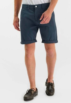 Uomo con pantaloncini blu navy risvoltati, scarpe stringate nere in pelle e una maglietta bianca, in piedi su sfondo bianco.