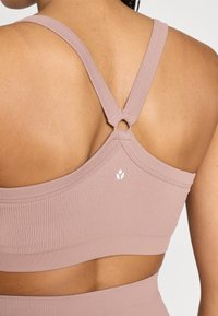 Ανοιχτό ροζ αθλητικό μπουστάκι με ριμπ ύφανση, φαρδιές τιράντες και κυκλικό μεταλλικό διακοσμητικό στη πίσω πλευρά. Διαθέτει μικρό λευκό λογότυπο στο πλάι.