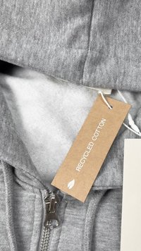 Siva kapuca izdelana iz recikliranega bombaža, s srebrnim zadrgo, mehkim notranjim oblogom in oznako, pritrjeno z navodili za nego.