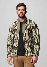 Fleecejacke im Tarnmuster mit Grüntönen und Beige, ausgestattet mit einem hohen Kragen und Reißverschluss, kombiniert mit einem schwarzen Hemd darunter.