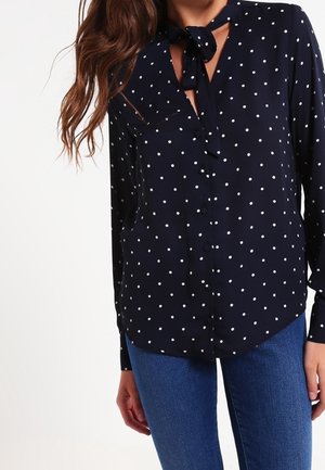 Vrouw draagt een marineblauwe blouse met lange mouwen, witte stippen en een strikdetail aan de hals, gecombineerd met een blauwe spijkerbroek.