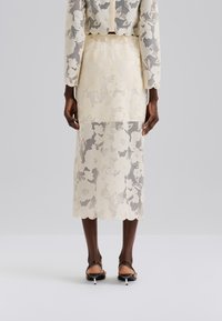 Spitzen-Zweiteiler mit Blumenmuster; durchscheinender, elfenbeinfarbener Stoff; wellenförmiger Saum am Rock; kombiniert mit braunen Sandalen mit Absatz.