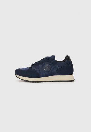 Navy blue athletic shoe with suede and mesh upper, round logo patch, lace-up design, cushioned sole, and rubber outsole.