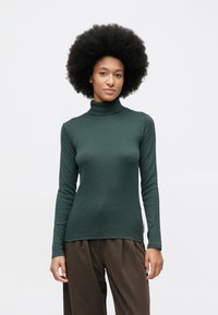 Junge Frau mit Afro-Frisur, die einen dunkelgrünen Rollkragenpullover und lockere braune Hosen trägt, steht vor einem schlichten weißen Hintergrund.