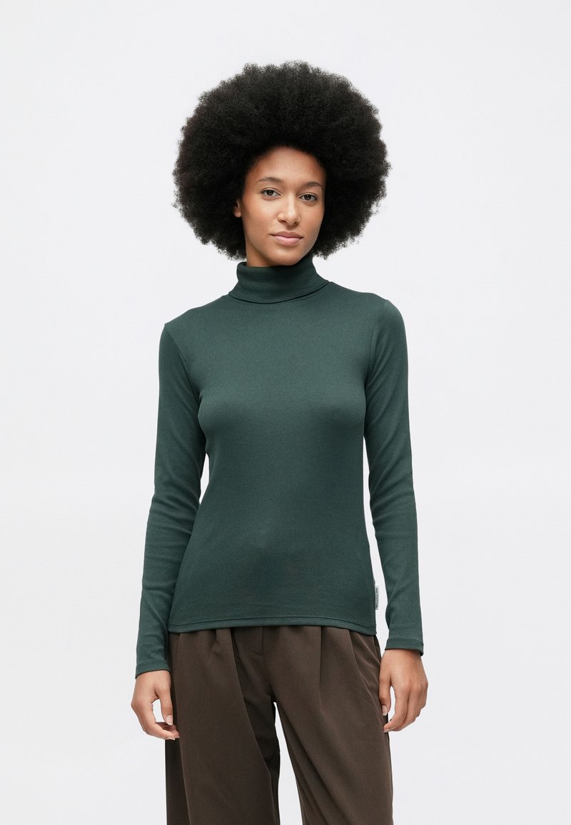 Junge Frau mit Afro-Frisur, die einen dunkelgrünen Rollkragenpullover und lockere braune Hosen trägt, steht vor einem schlichten weißen Hintergrund.