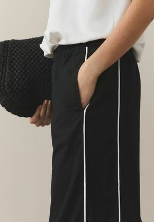 Osoba ubrana w czarne spodnie z białymi paskami po bokach, biała koszula włożona do środka, ręka w kieszeni, trzymająca czarną plecioną torbę.