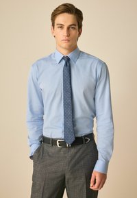 Helles blaues Hemd mit Button-Down-Kragen, getragen mit einer dunkelblauen Krawatte mit Punkten und grauen strukturierten Hosen, gehalten von einem schwarzen Gürtel.