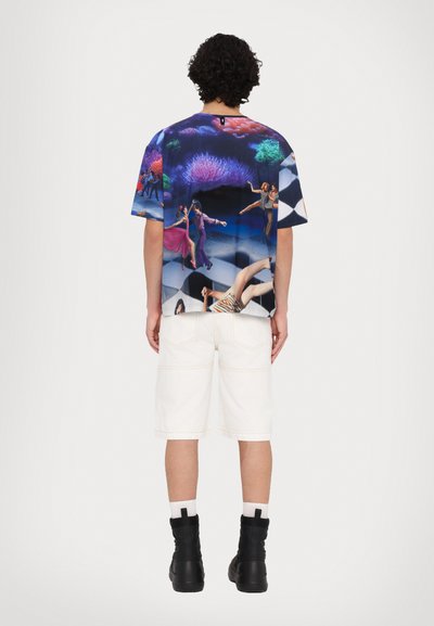 Camicia a maniche corte con un design grafico colorato che presenta danzatori, motivo a scacchi in bianco e nero, indossata con pantaloni corti bianchi e stivali neri.