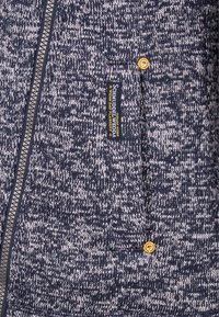 Marinblå och grå texturerad zip-up hoodie med en tygetikett och en liten gul knappdetalj på framfickan.