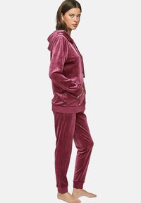 Ensemble de survêtement en velours de couleur rose profond, comprenant un haut zippé à capuche avec poches et un pantalon fuselé assorti avec des poignets élastiques. Texture lisse.