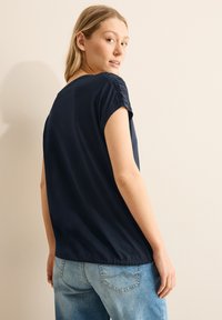 Top de manga corta azul marino con una textura suave, que presenta detalles recogidos en los hombros y un ajuste relajado, combinado con unos jeans de denim claro.