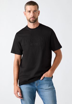Mand med kort hår og skæg iført en sort Guess t-shirt og blå jeans, stående med den ene hånd i lommen mod en ensfarvet baggrund.