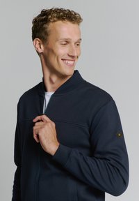 Marineblauw zippered jack met een gestructureerd oppervlak, staande kraag en zijzakken. Bevat een badge op de linker mouw.