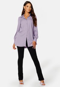 Lawendowa koszula zapinana na guziki z długimi rękawami i marszczonymi mankietami, zestawiona z czarnymi dopasowanymi spodniami i jasnofioletowymi sandałami na wysokim obcasie.
