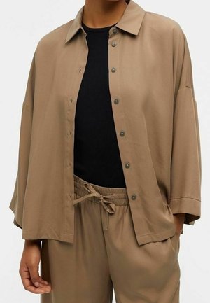 Personne portant une chemise beige ample boutonnée sur un top noir et un pantalon beige à cordon assorti, les mains dans les poches.