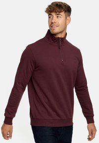 Burgunderfarbener Reißverschluss-Pullover aus strukturiertem Stoff. Verfügt über einen hohen Kragen, lange Ärmel und ein dezentes Logo auf der Brust.