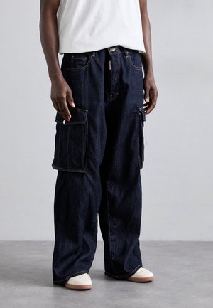Dark denim cargo pants with wide legs, two large side pockets, yellow stitching, and a button closure at the waist. Worn with light sneakers.