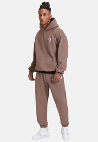 Felpa in pile marrone con tasca frontale e patch con logo, abbinata a pantaloni jogger coordinati con polsini elasticizzati e sneakers bianche.