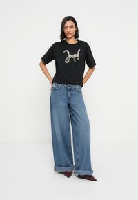 Tricou negru cu text grafic metalic, asortat cu blugi largi de culoare albastru deschis, cu tivuri rulate și pantofi burgundy cu vârf ascuțit.