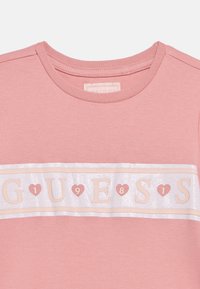 Ροζ βαμβακερό T-shirt με στρογγυλή λαιμόκοψη, που διαθέτει λευκή και απαλό ροζ λωρίδα με την επιγραφή "GUESS 1981" με κεφαλαία γράμματα και χρωματικές καρδιές.