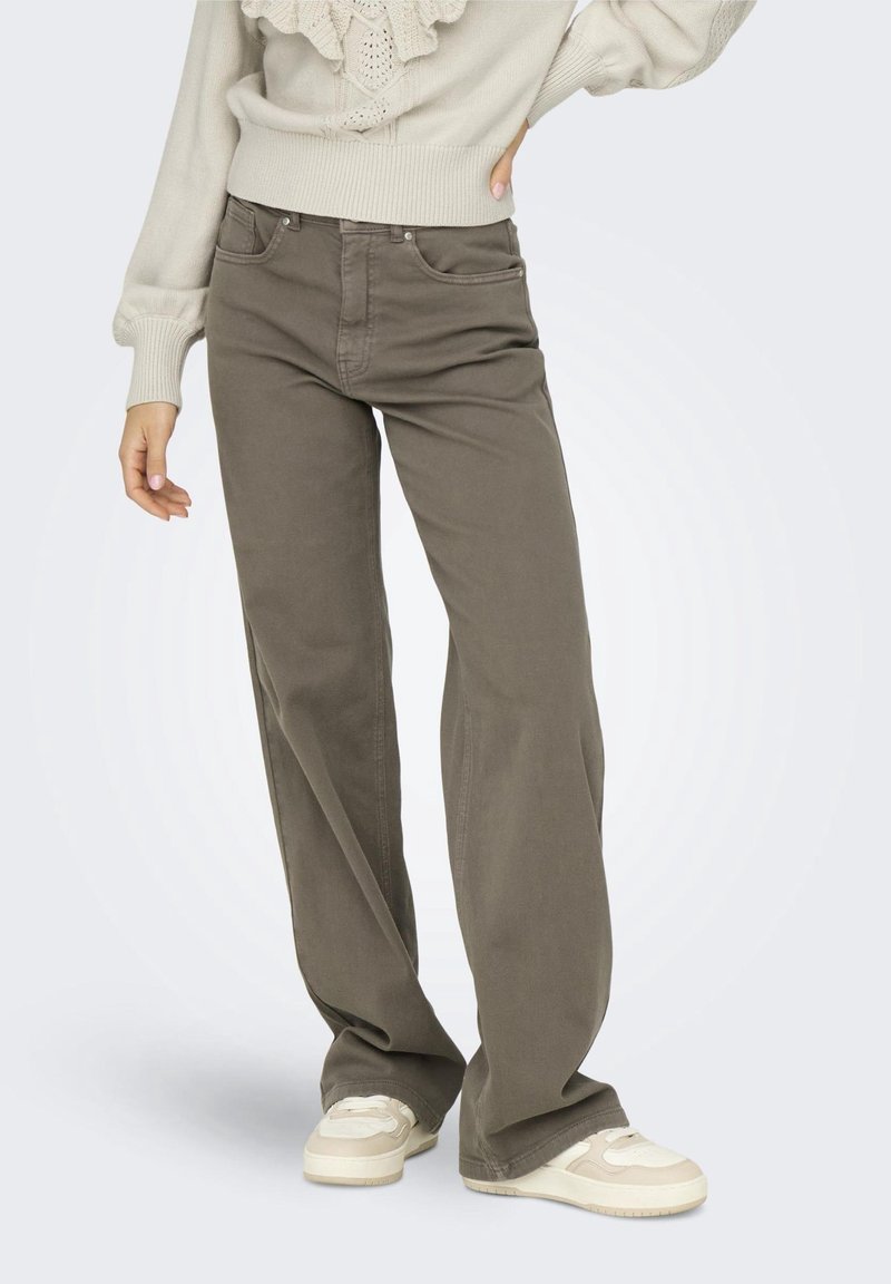 Personne portant un pantalon large taille haute marron avec des poches avant, associé à des baskets blanches et un pull en tricot crème.