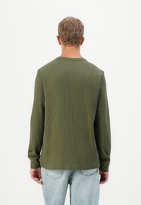 Mann som står med ryggen til kameraet iført en olivengrønn ribbestrikket langermet skjorte og lyseblå denimjeans mot en hvit bakgrunn.
