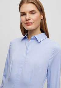 Hele halli pikkade varrukatega särk, millel on terav krae, sile tekstuur, esine nupulukk ja õrn õmblusdetail rinnal.